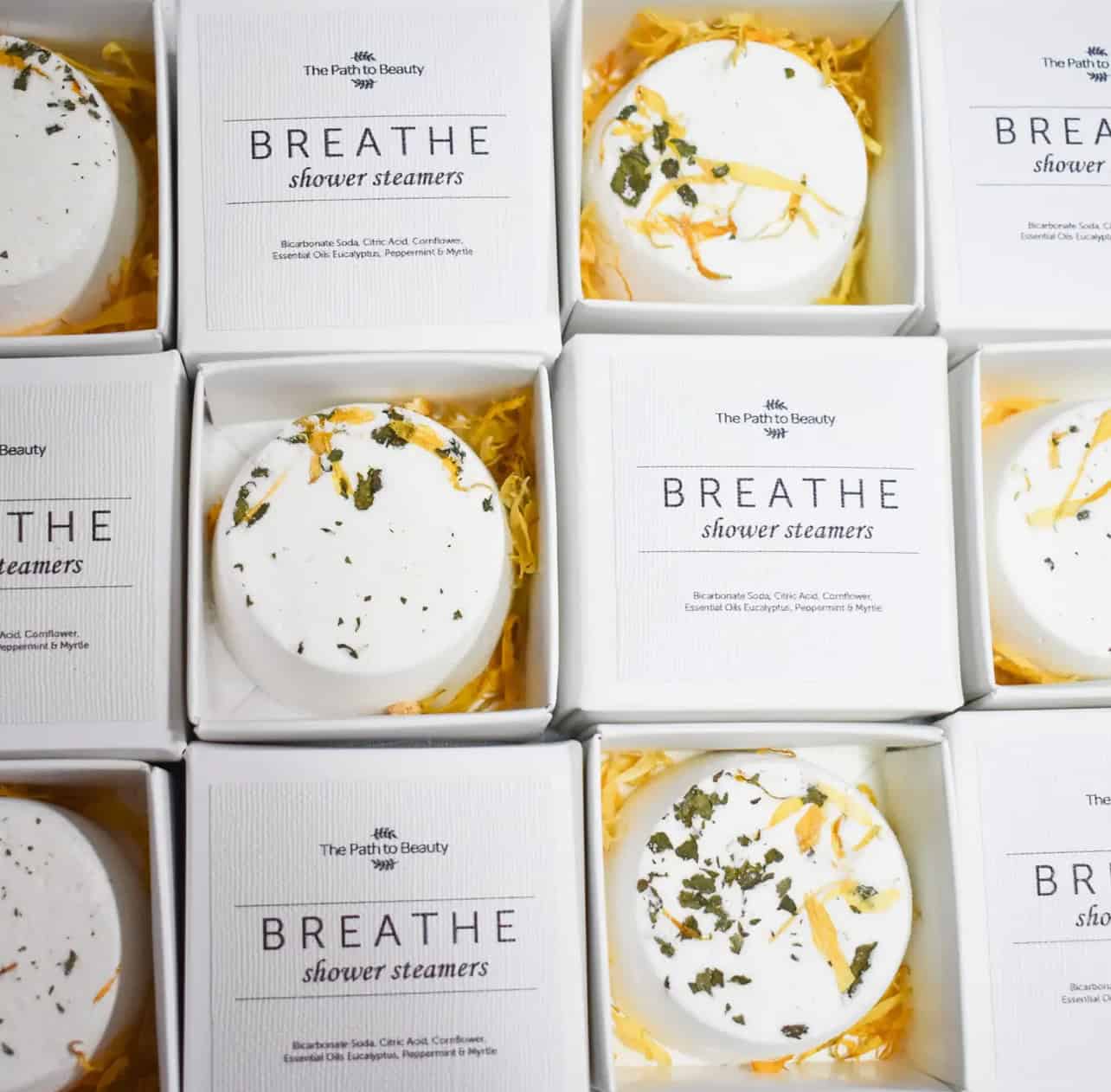Nine boxes of Breathe Individual Shower Steamers are arranged in a grid. Each round, white steamer with dried herbs rests in yellow shredded paper inside its labeled box.