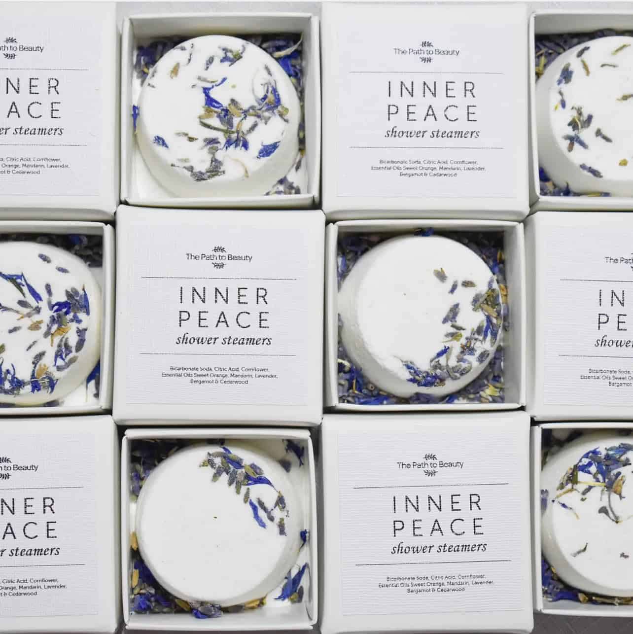 A grid of boxed Inner Peace Individual Shower Steamers—each round, white tablet topped with dried blue and yellow petals—displayed neatly in white packaging.