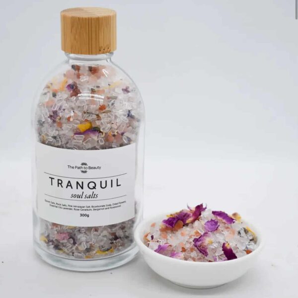 A clear glass bottle labeled Tranquil Bath Salts holds colorful salts, sitting beside a small white bowl with a portion of the mixture, featuring visible flower petals and crystals for ultimate relaxation.