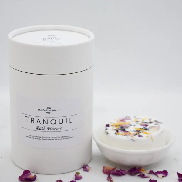 A white cylindrical container labeled Bath Fizzers is placed beside a round bath fizzer with dried flower petals on top, both displayed against a simple white background.