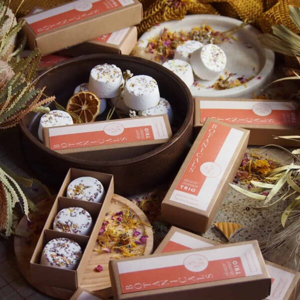 Botanicals Shower Steamers, both boxed and unboxed, are displayed on wooden trays with white bath bombs and topped with dried flowers, surrounded by botanicals, dried leaves, and petals.