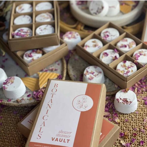 Boxes of round, white Geranium Lavender Shower Steamers, infused with floral scents and topped with dried petals, are scattered on a woven surface. One box is labeled Botanicals Shower Steamer Vault.