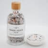 A clear glass bottle labeled Inner Peace Bath Salts with a wooden lid is placed beside a small white bowl filled with these colorful bath salts, which feature mixed minerals and dried flower petals.