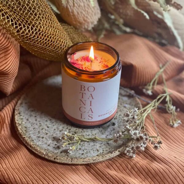 The Pear & Cinnamon Botanical Candle in an amber glass jar sits on a speckled ceramic plate, surrounded by dried flowers and textured brown fabric, creating a warm and cozy ambiance.