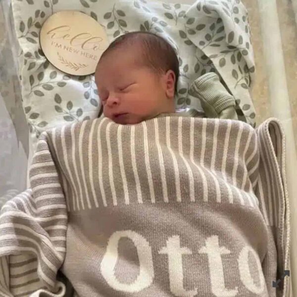 A newborn sleeps peacefully in a cozy beige and white Custom Personalised & Photo Blanket with the name Otto. A round wooden sign says Hello, Im new here, set against a soft background of green leafy fabric.