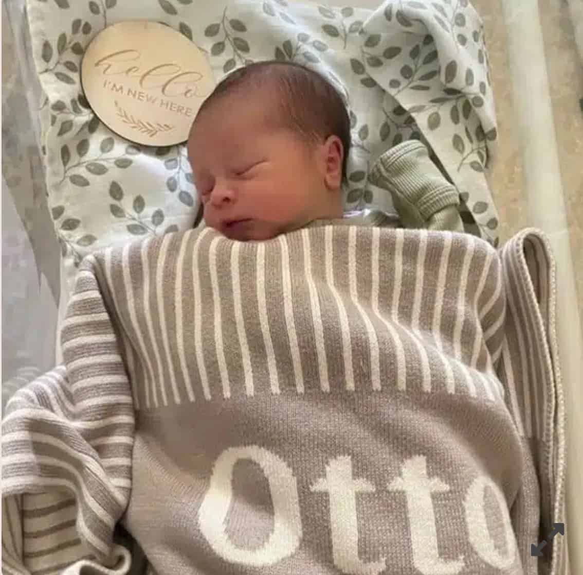 A newborn sleeps peacefully in a cozy beige and white Custom Personalised & Photo Blanket with the name Otto. A round wooden sign says Hello, Im new here, set against a soft background of green leafy fabric.