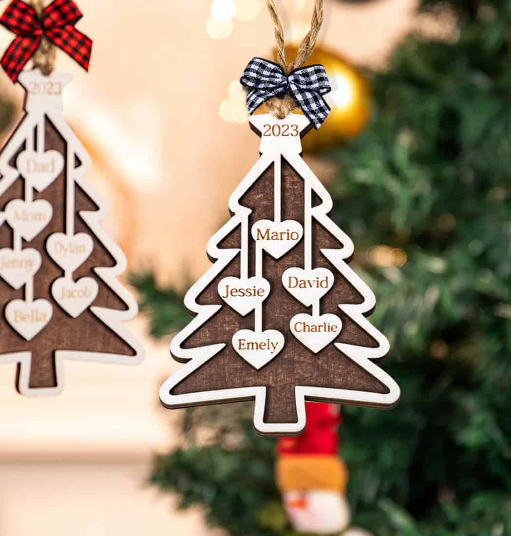 The Custom Christmas Tree Ornament hangs on a tree, featuring heart-shaped spaces labeled Mario, Jessie, David, Emely, and Charlie with a black-and-white bow and 2023 displayed at the top.