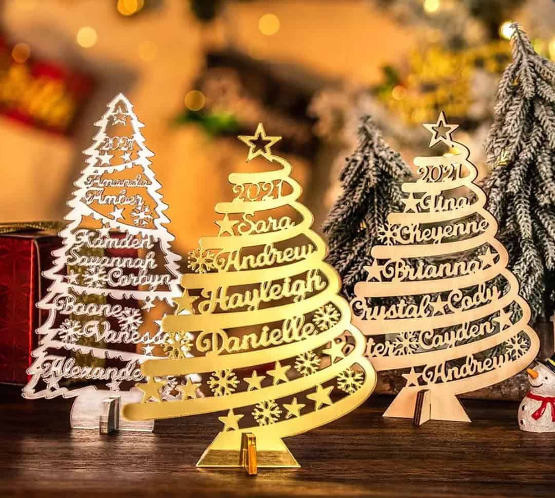 Three Personalised Christmas Tree ornaments in Gold, Silver, or Wood, each with custom names and the year 2021, arranged on a festive table with blurred lights and mini trees in the background.