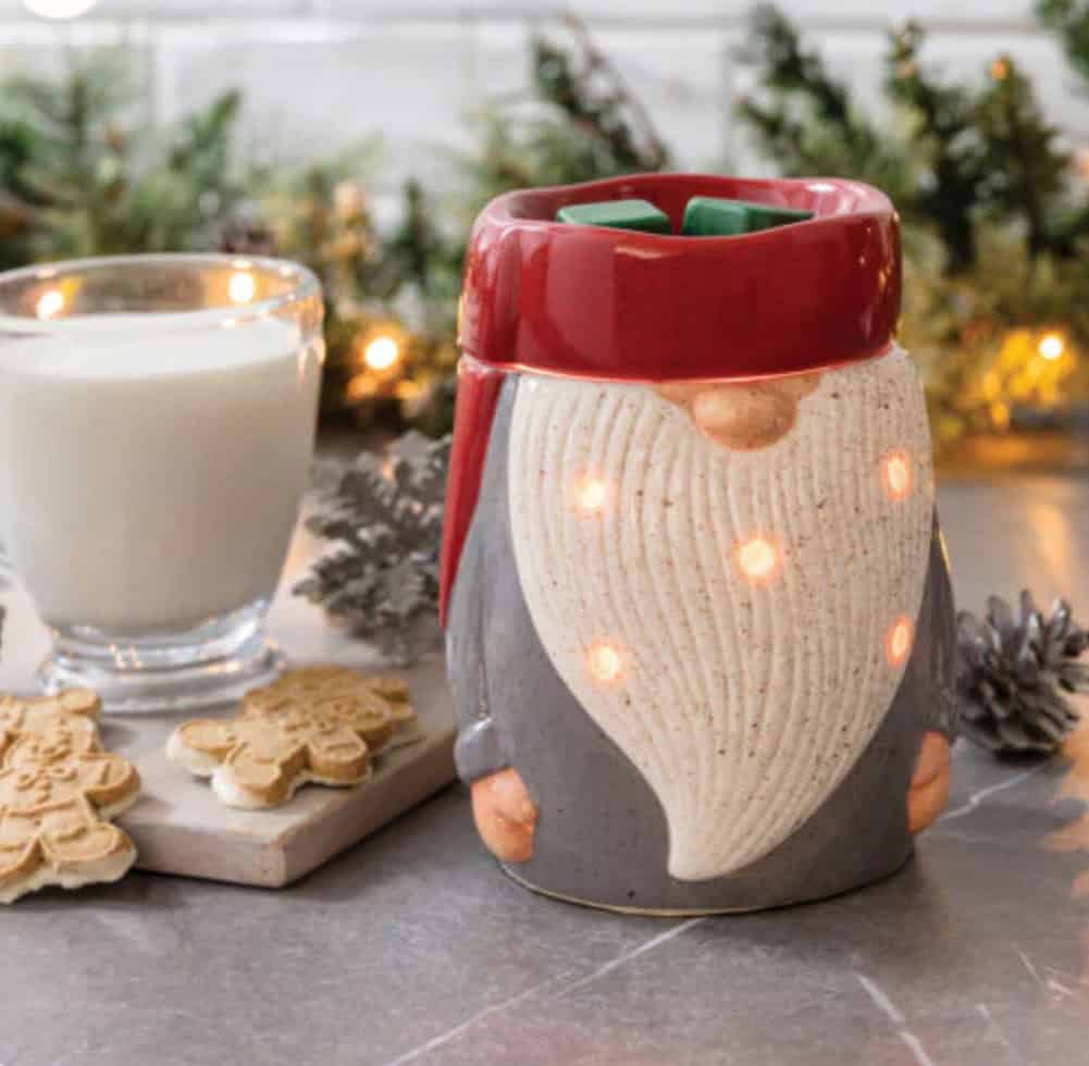 The Gnome Wax Warmer with glowing lights sits on a counter beside a glass of milk, pinecones, decorated gingerbread cookies, and festive greenery in the background.