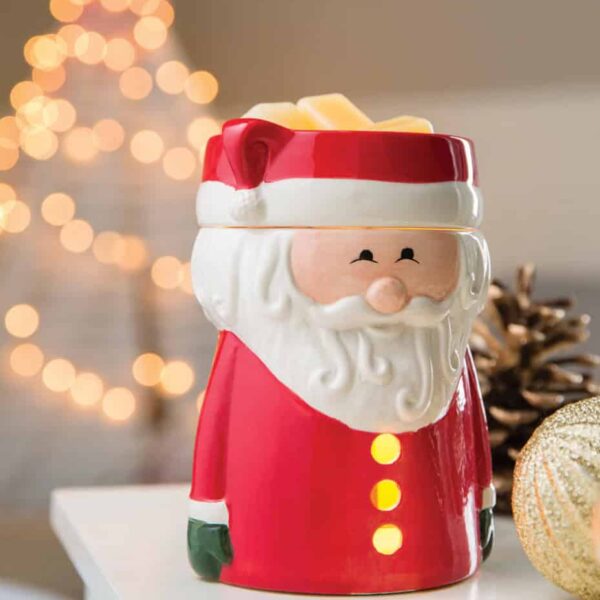 A Santa Claus Wax Warmer sits on a white surface. In the background, blurred holiday lights, a gold ornament, and a pinecone evoke the cozy spirit of Christmas.