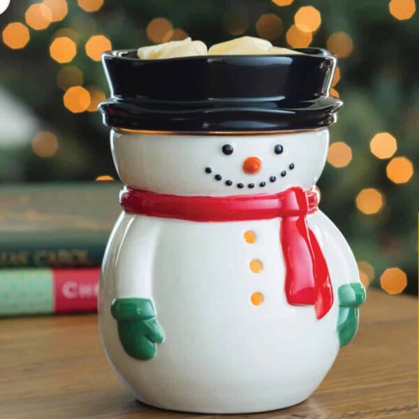 A Frosty Snowman Wax Warmer with a red scarf, green mittens, and black hat sits on a wooden table. The background shows blurred holiday lights and books.