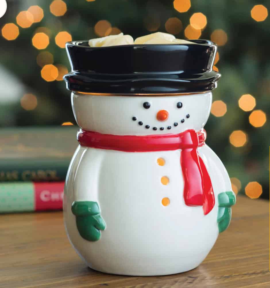 A Frosty Snowman Wax Warmer with a red scarf, green mittens, and black hat sits on a wooden table. The background shows blurred holiday lights and books.