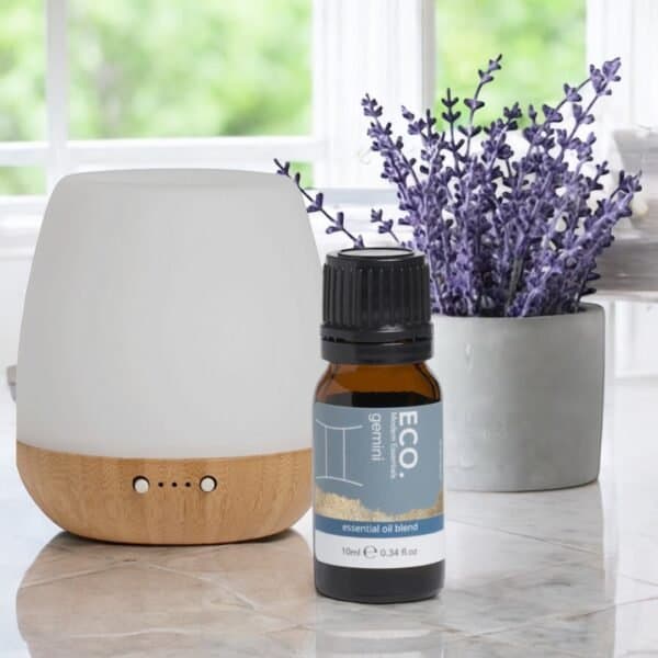 A white and wood essential oil diffuser sits beside a small bottle of Gemini Zodiac Essential Oil Blend, ideal for Geminis. Behind them, a pot of lavender flowers is placed on a windowsill overlooking greenery.