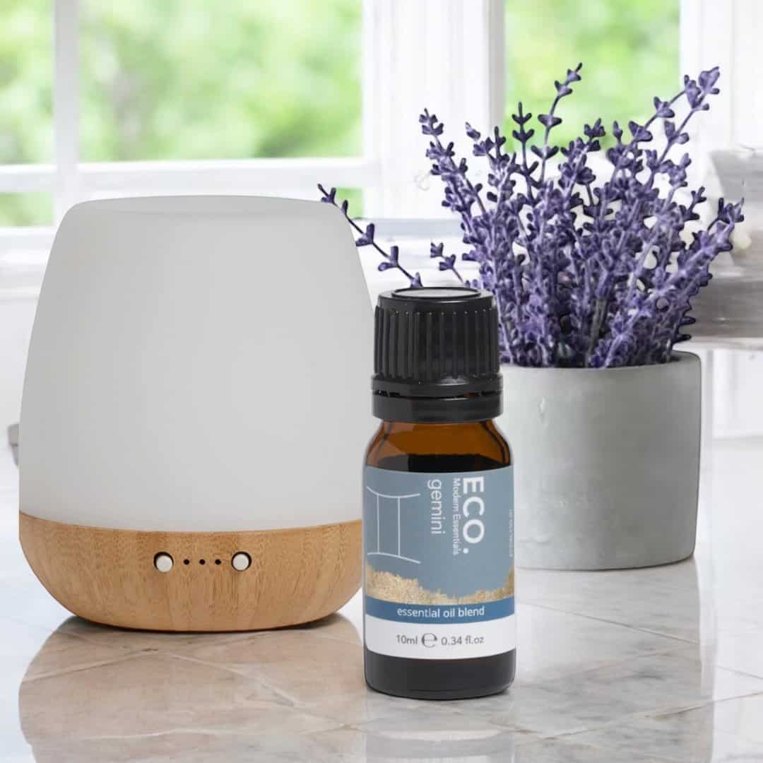 A white and wood essential oil diffuser sits beside a small bottle of Gemini Zodiac Essential Oil Blend, ideal for Geminis. Behind them, a pot of lavender flowers is placed on a windowsill overlooking greenery.