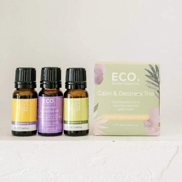 Three bottles of essential oils—Lavender, Lemongrass, and Lemon—stand beside a pastel green Calm and De-Stress Essential Oil Trio Pack box on a white surface, ideal for anyone seeking relaxation and stress relief.