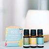 Three ECO. essential oil bottles stand before a pastel Sunshine State of Mind Essential Oil Blends Trio Pack box, with a white diffuser blurred in the background—perfect for uplifting aromatherapy sessions.
