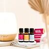 Three bottles from the Rest and Glow Essential Oil Trio Pack sit on a coaster beside a white diffuser, folded cloth, and pink eco-branded box. Soft lighting adds to the serene atmosphere.