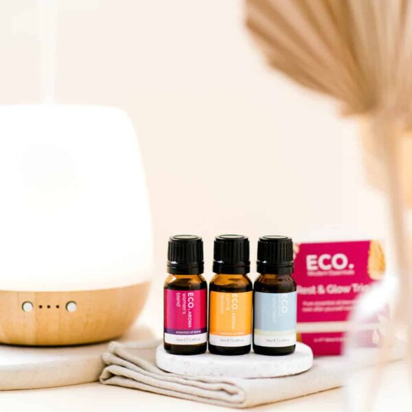 Three bottles from the Rest and Glow Essential Oil Trio Pack sit on a coaster beside a white diffuser, folded cloth, and pink eco-branded box. Soft lighting adds to the serene atmosphere.