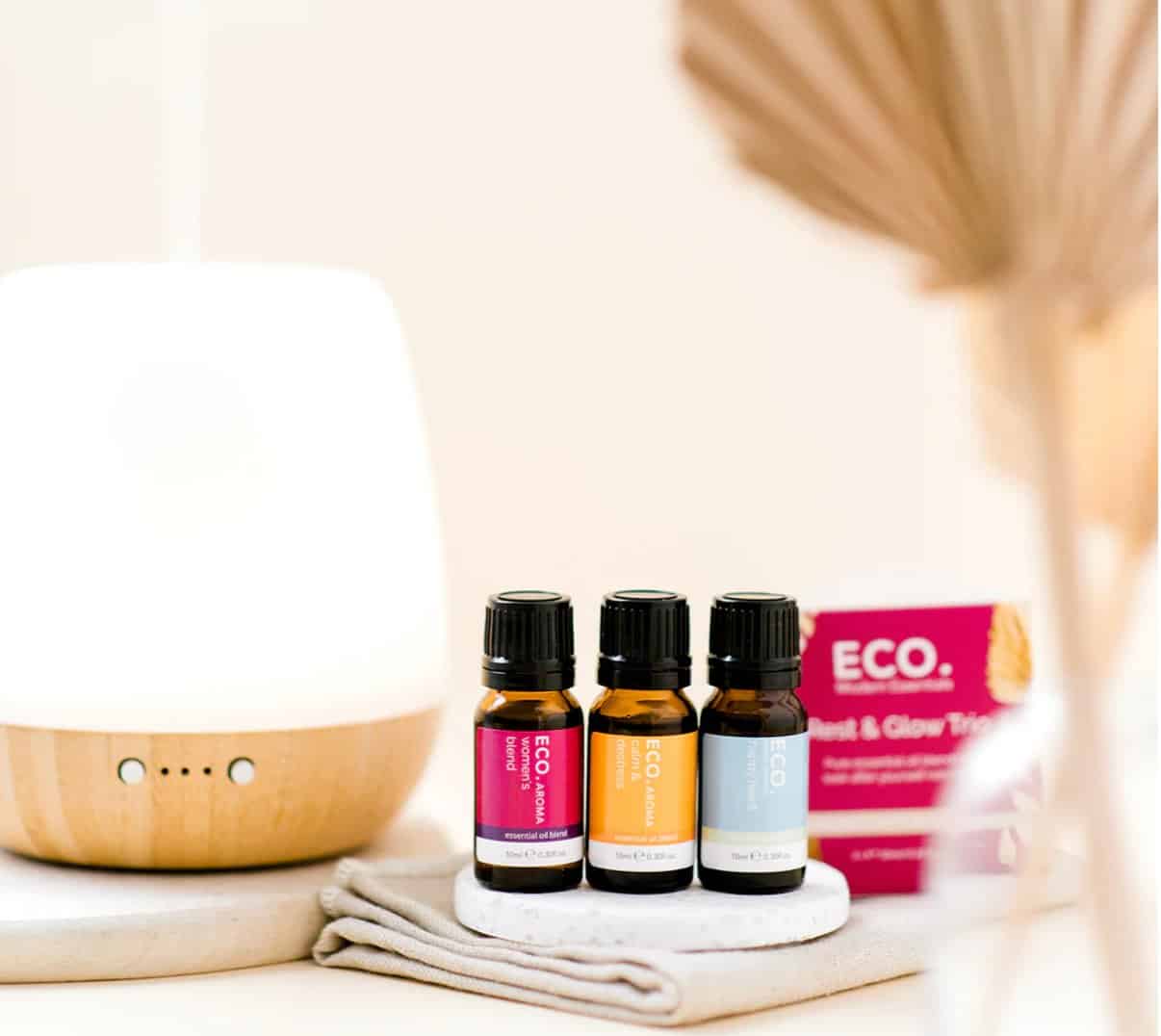 Three bottles from the Rest and Glow Essential Oil Trio Pack sit on a coaster beside a white diffuser, folded cloth, and pink eco-branded box. Soft lighting adds to the serene atmosphere.