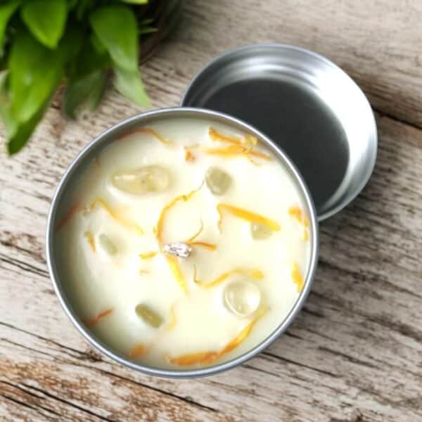 The Prosperity & Abundance Travel Tin Candle, filled with white wax and adorned with yellow flower petals and translucent stones, sits on a wooden surface near a green leafy plant. Its open lid rests beside the tin.
