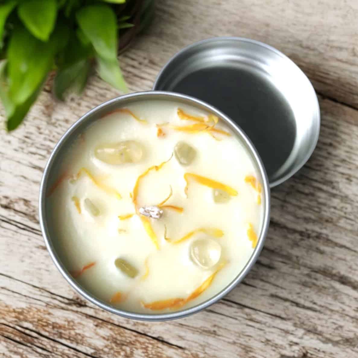 The Prosperity & Abundance Travel Tin Candle, filled with white wax and adorned with yellow flower petals and translucent stones, sits on a wooden surface near a green leafy plant. Its open lid rests beside the tin.