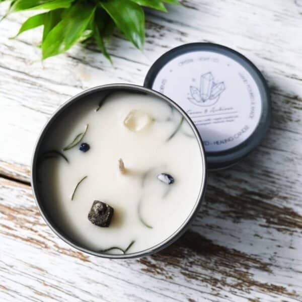 A Focus & Achieve Crystal Healing Candle in a metal tin with crystals and herbs in the wax sits on a rustic white wooden surface, a green plant nearby. The labeled lid rests beside the candle.