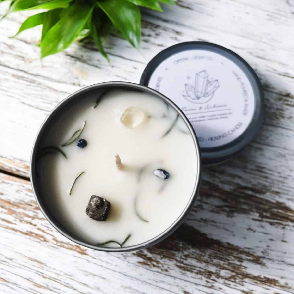 A Focus & Achieve Crystal Healing Candle in a metal tin with crystals and herbs in the wax sits on a rustic white wooden surface, a green plant nearby. The labeled lid rests beside the candle.