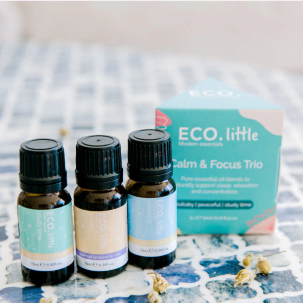 Three small essential oil bottles stand before a blue and teal ECO Little Calm and Focus box on a blue-tiled surface, with dried flowers scattered nearby for a serene touch.