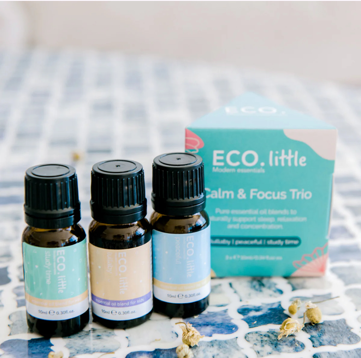 Three small essential oil bottles stand before a blue and teal ECO Little Calm and Focus box on a blue-tiled surface, with dried flowers scattered nearby for a serene touch.