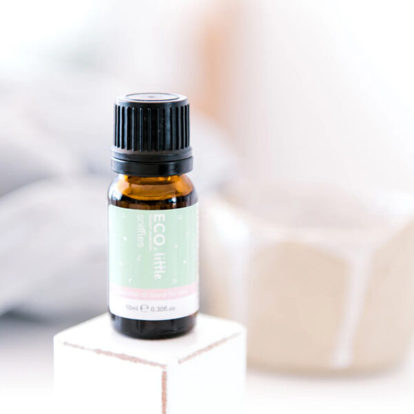A 10ml (0.30 fl oz) amber bottle of ECO Little Sniffles Essential Oil Blend rests on a white block, with a blurred beige cup in the soft background.