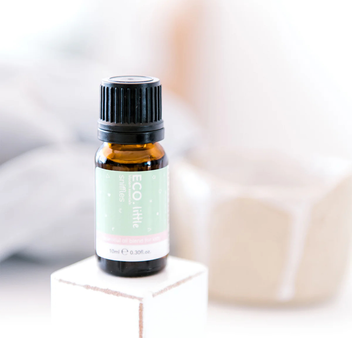 A 10ml (0.30 fl oz) amber bottle of ECO Little Sniffles Essential Oil Blend rests on a white block, with a blurred beige cup in the soft background.