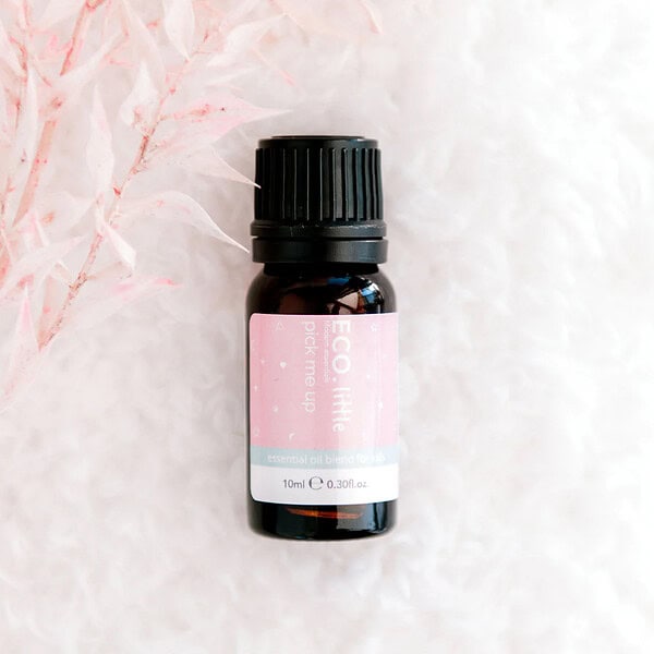 A 10ml bottle of ECO Little Pick Me Up Essential Oil Blend sits on a soft white textured surface, accented by delicate pink-tinted foliage on the left.