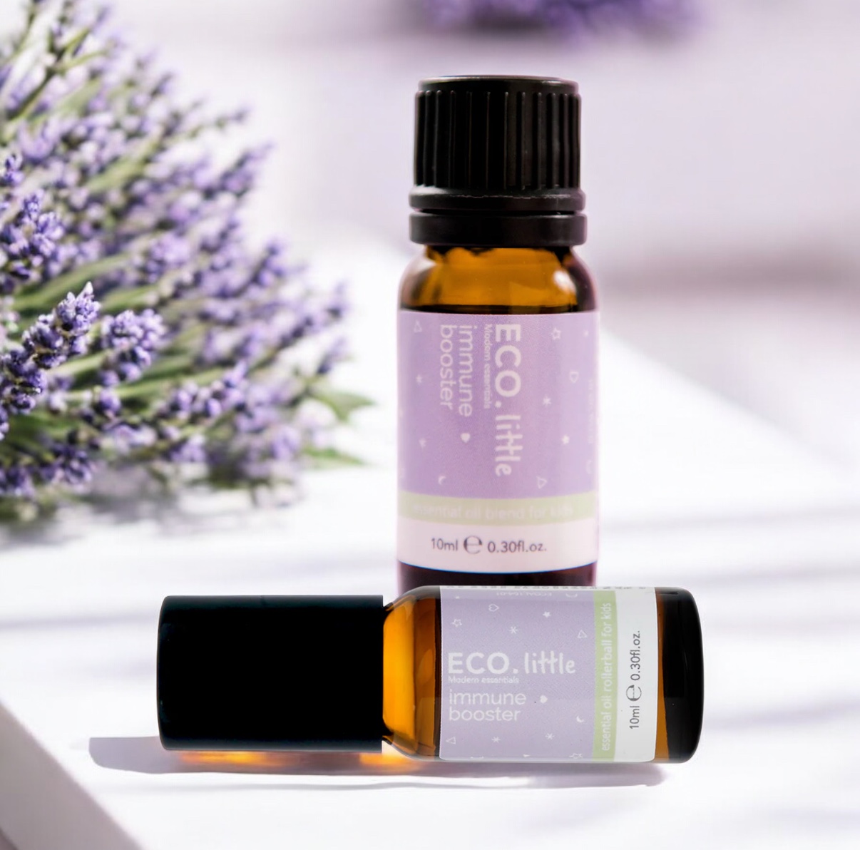 Two brown glass bottles of ECO Little Immune Booster Essential Oil or Roller Blend sit on a white surface, with lavender sprigs blurred in the background. One bottle stands upright; the other roller blend lies horizontally in front.