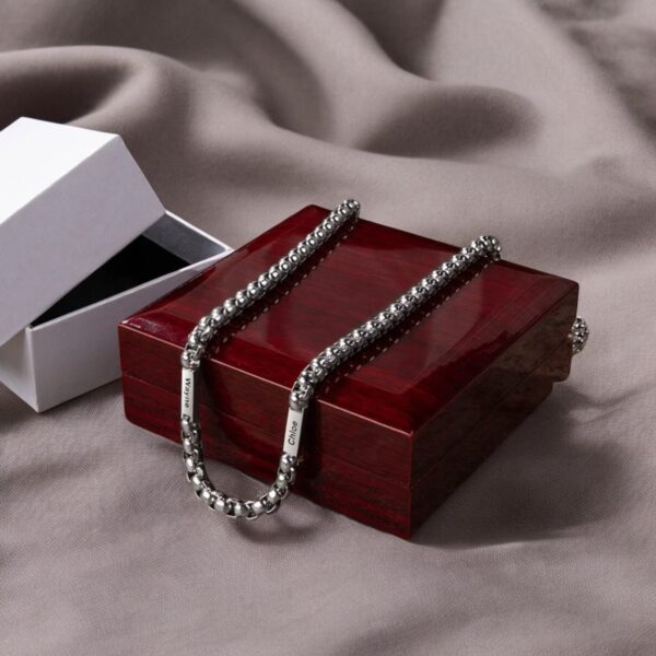 The Personalised 2 Name Cuban Chain Necklace For Him is displayed on a polished wooden jewelry box, with its white gift box lid open nearby, all set against a soft gray fabric background.