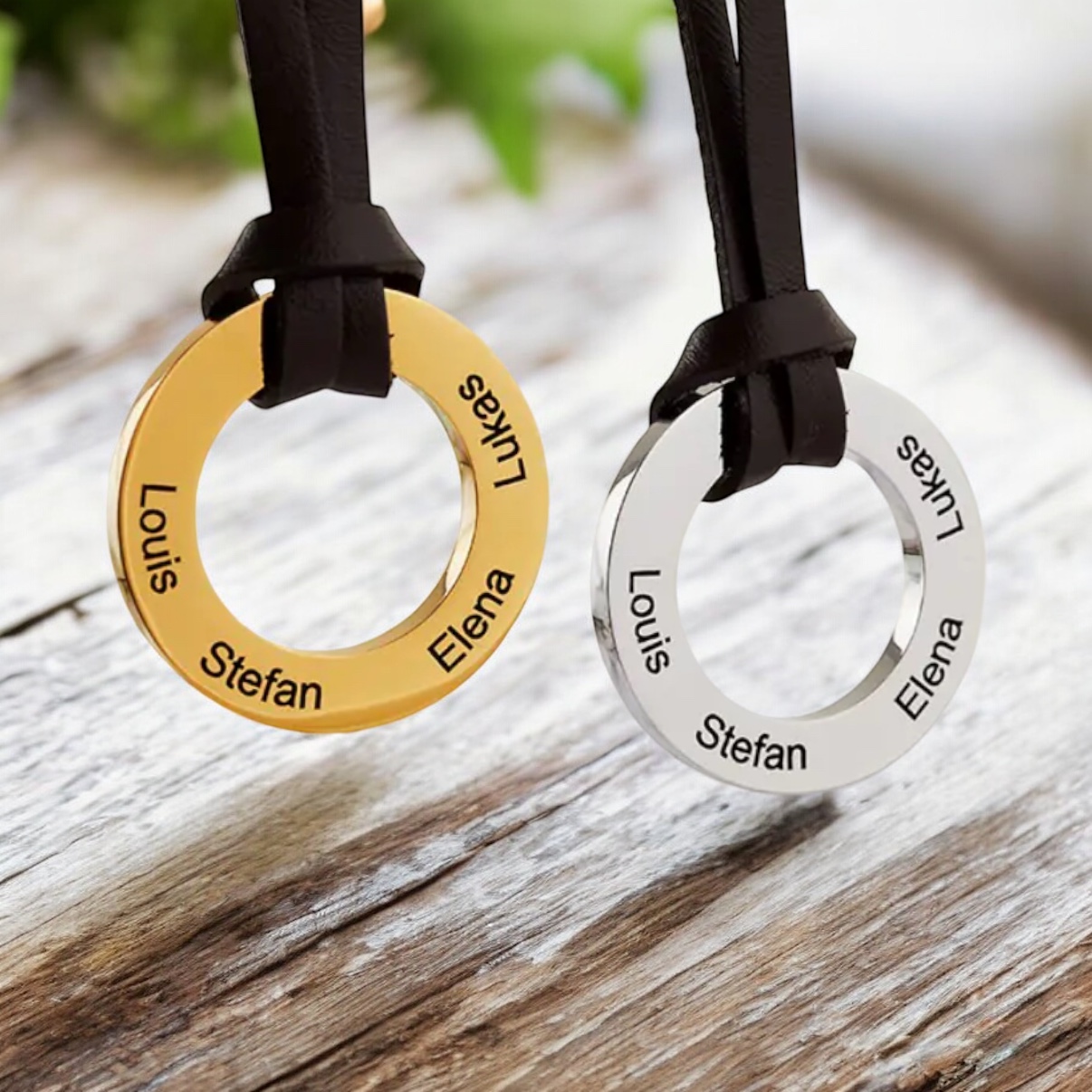 Two Personalised Circle Pendant Necklace on a Leather Strap—one gold, one silver—engraved with Louis, Stefan, Elena, and Lukas rest elegantly on a rustic wooden surface.