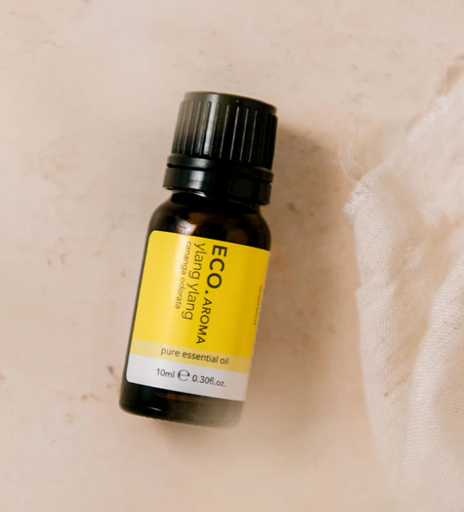 A small dark amber bottle labeled Ylang Ylang Pure Essential Oil sits on a light surface beside a piece of white fabric.