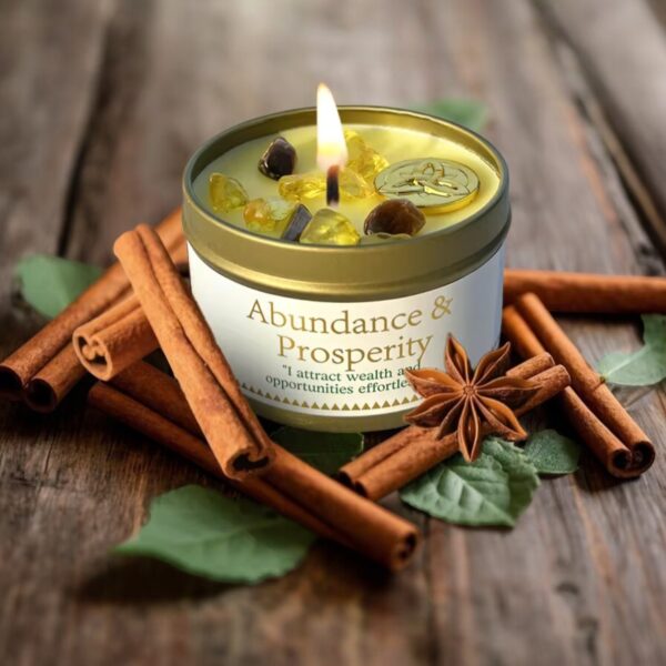 An Abundance Intention Candle - Cinnamon, Patchouli, Citrine, Pyrite tin candle in a gold tin sits on wood, surrounded by cinnamon sticks, star anise, and green leaves. The candle features citrine stones and a gold coin to inspire abundance.