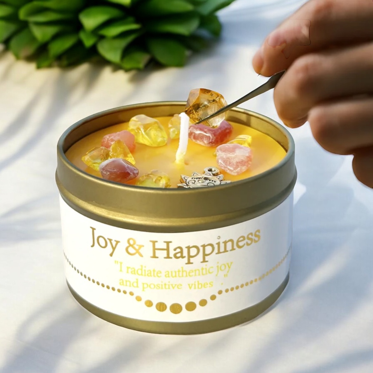 A hand places decorative stones onto a yellow candle in a tin labeled Joy & Happiness with the phrase I radiate authentic joy and positive vibes. A green succulent is blurred in the background.