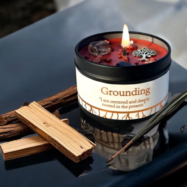 A lit Grounding Intention Candle - Cedarwood & Vetiver, labeled Grounding and inscribed with I am centered and deeply rooted in the present, sits on a dark surface surrounded by wood sticks, a feather, and decorative stones.