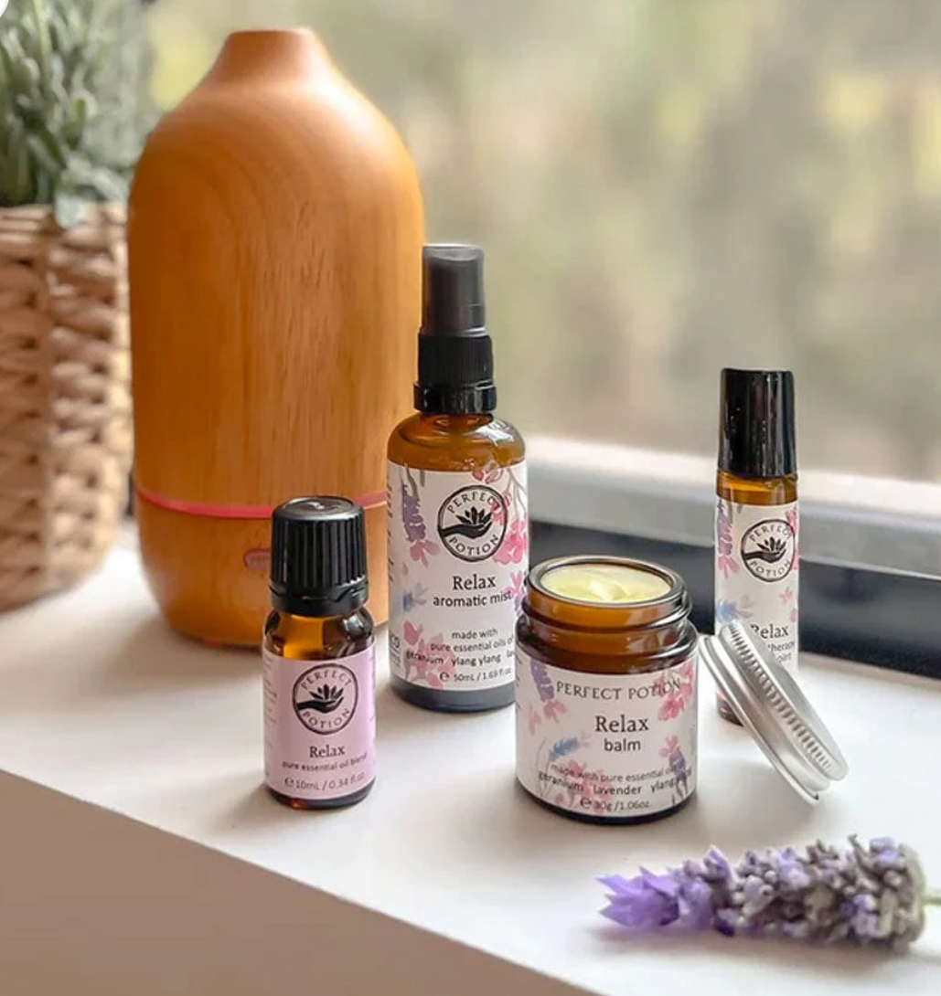The Perfect Potion Relax Balm is displayed on a table with a sprig of lavender, a wooden diffuser, and a woven basket in the background.