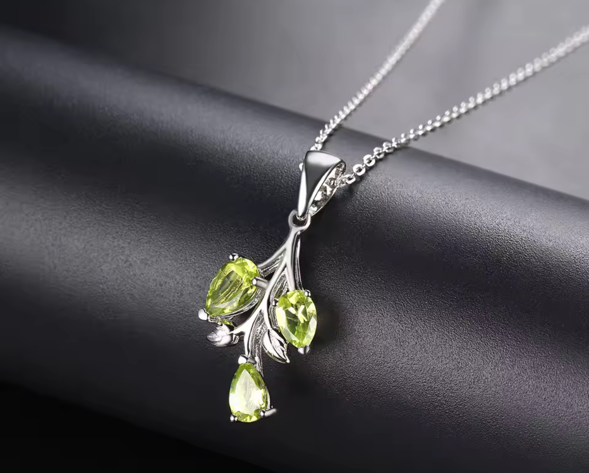 The Peridot 3 Tier Sterling Silver Pendant Necklace features a branch-shaped pendant with three green teardrop peridot gemstones, displayed on a matte black surface.