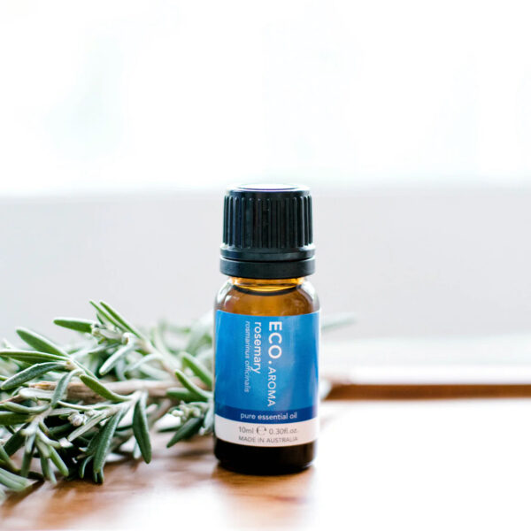 A small brown bottle of Rosemary Pure Essential Oil rests on a white surface beside fresh rosemary sprigs, set against a bright, blurred background.