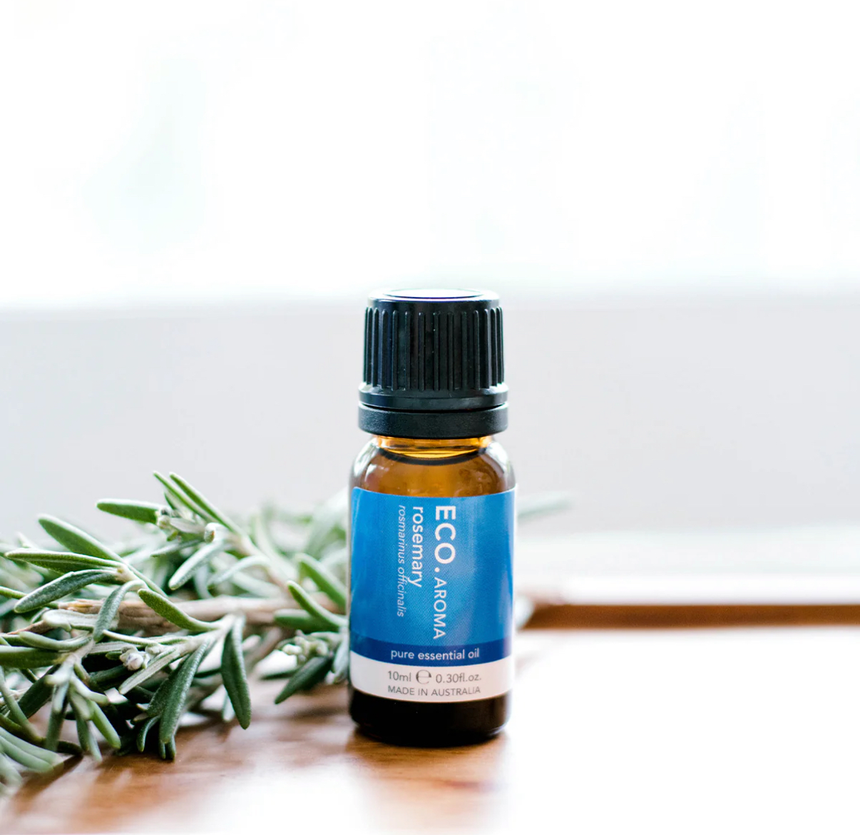 A small brown bottle of Rosemary Pure Essential Oil rests on a white surface beside fresh rosemary sprigs, set against a bright, blurred background.