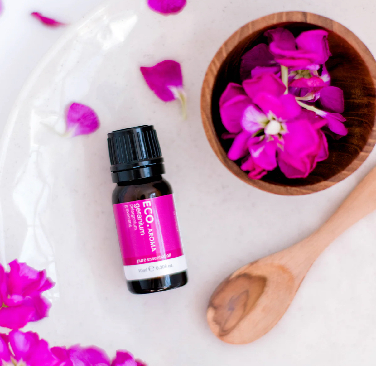 A bottle of Geranium Pure Essential Oil rests on a white surface beside a wooden bowl of pink flower petals and a wooden spoon, with scattered petals adding a vibrant touch to the scene.