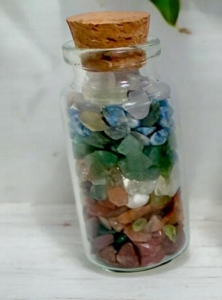 A small glass bottle with a cork stopper, filled with various colorful polished stones and crystals, arranged in layers. The bottle is placed on a white surface.