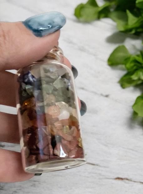 A hand with blue-tipped nails holds a small glass vial filled with layered, colorful gemstone chips. Out-of-focus green leaves rest on a light wooden surface in the background.