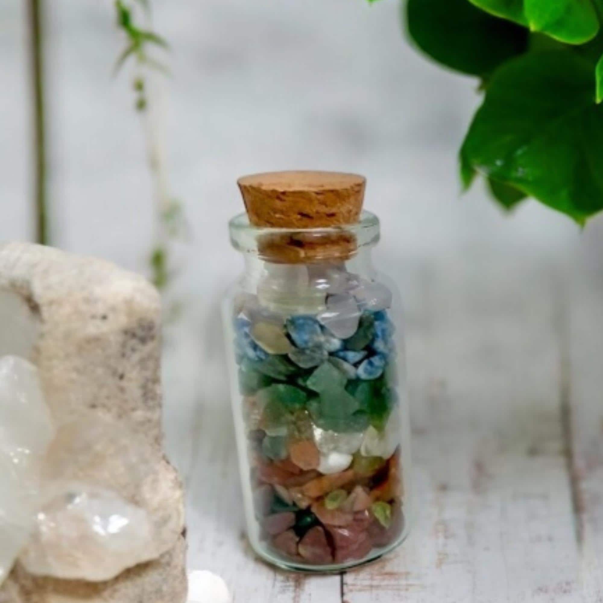 A small glass jar with a cork lid filled with colorful crystal chips sits on a white wooden surface next to a stone and green leafy plant.