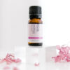 A small brown bottle of Jasmine Essential Oil rests on a clear acrylic block, surrounded by pink dried flower petals against a white background.