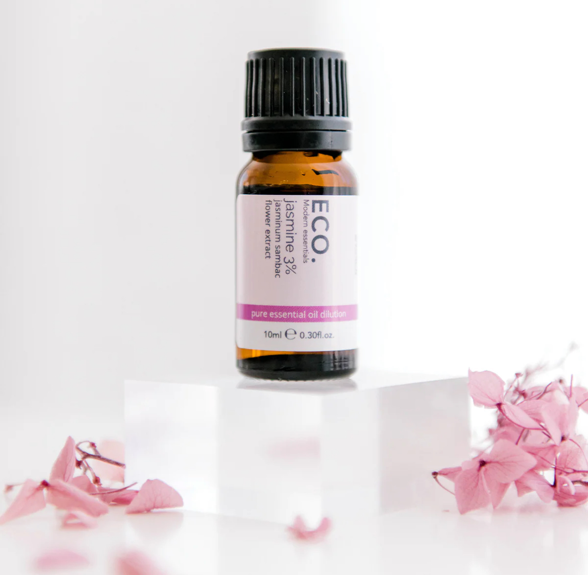 A small brown bottle of Jasmine Essential Oil rests on a clear acrylic block, surrounded by pink dried flower petals against a white background.