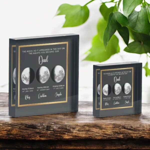Two acrylic photo blocks display images of the moon’s phases on specific dates, labeled with names and descriptions. The blocks are set on a wooden surface next to green leafy plants.